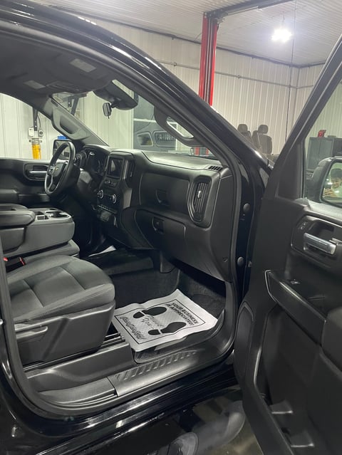 Interior of black pickup truck with open driver-side door, steering wheel, dashboard, and gray seats in warehouse setting