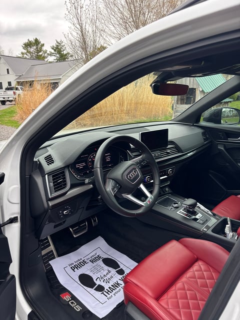 Interior view of an Audi sports car with red leather seats, steering wheel, and dashboard, window open to residential driveway